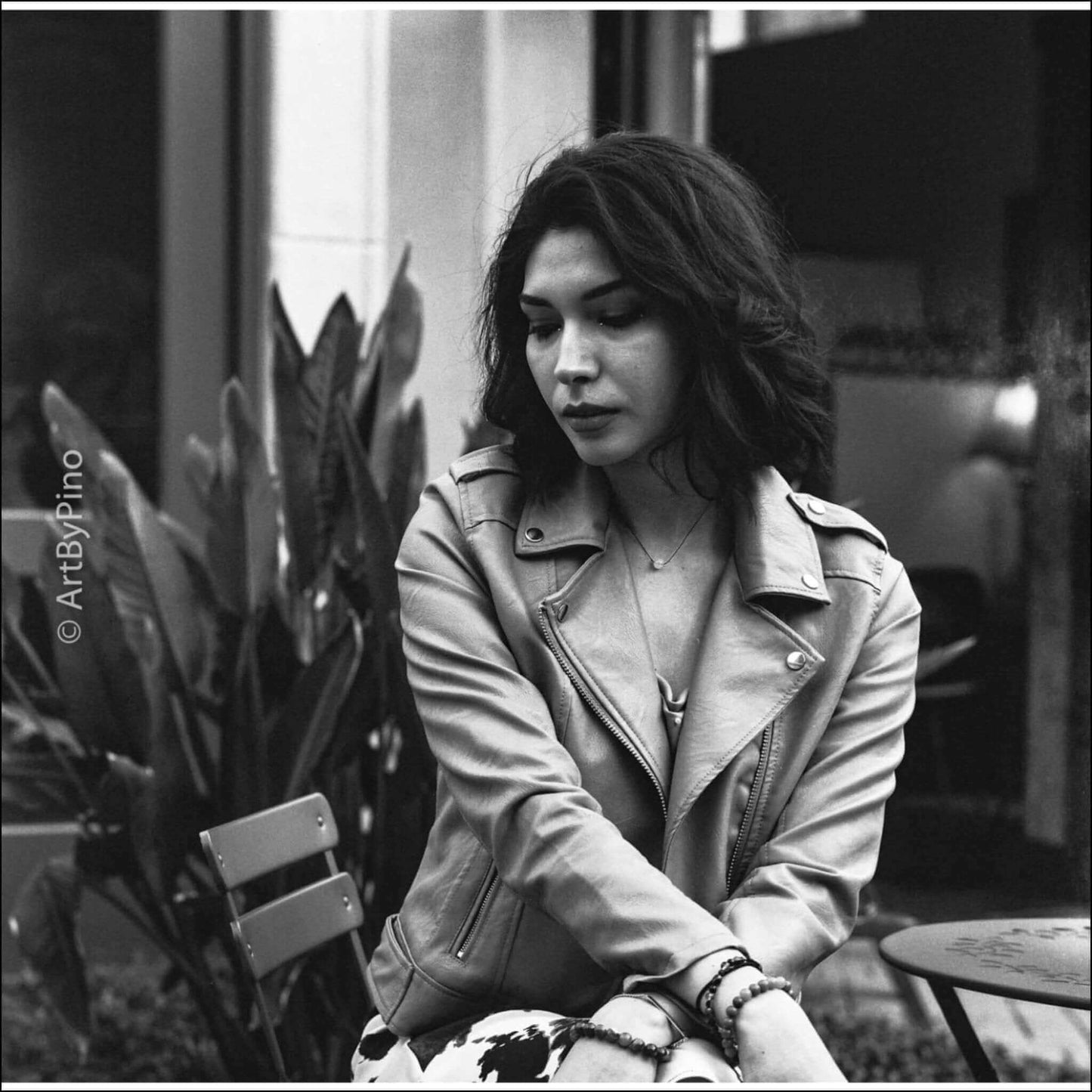 A contemplative woman in a leather jacket sitting outdoors among greenery.