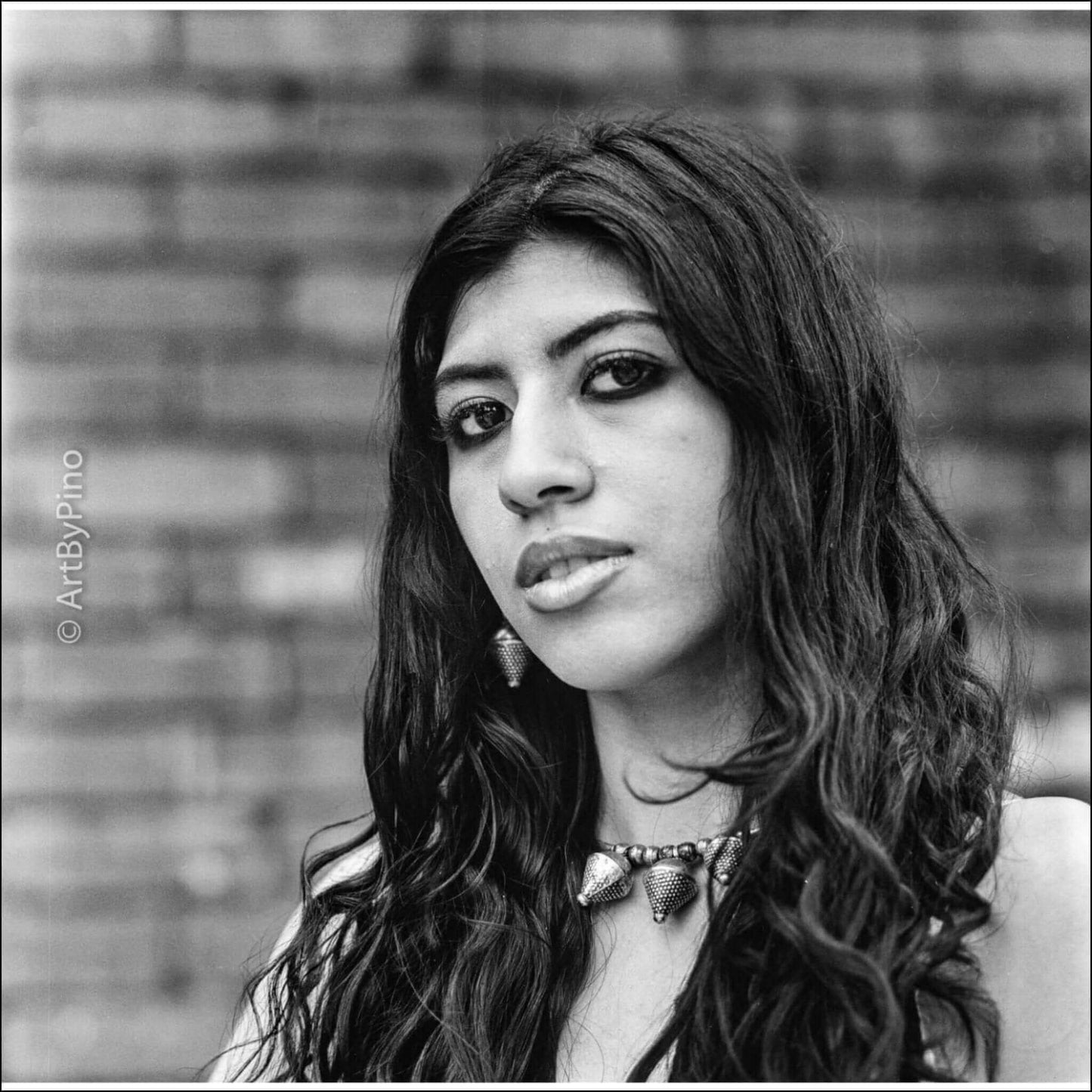 Black and white portrait of a woman with long hair, wearing earrings and a necklace, against a textured brick background.