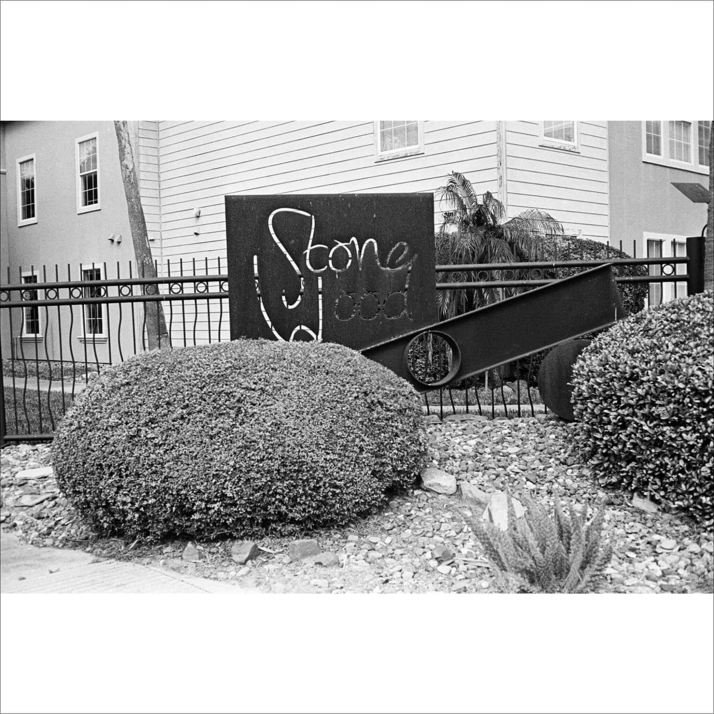 Black-and-white photo of a sign reading 'Stone Wood' next to landscaped bushes and a fence.
