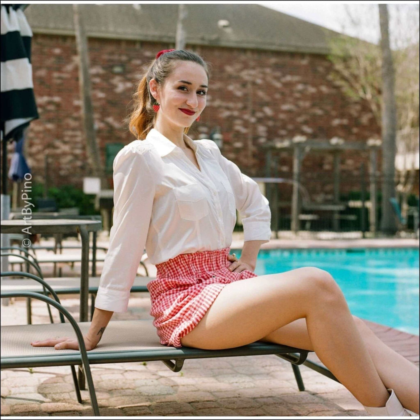 Woman in a white shirt and red checkered shorts sitting by a pool, enjoying a sunny day.
