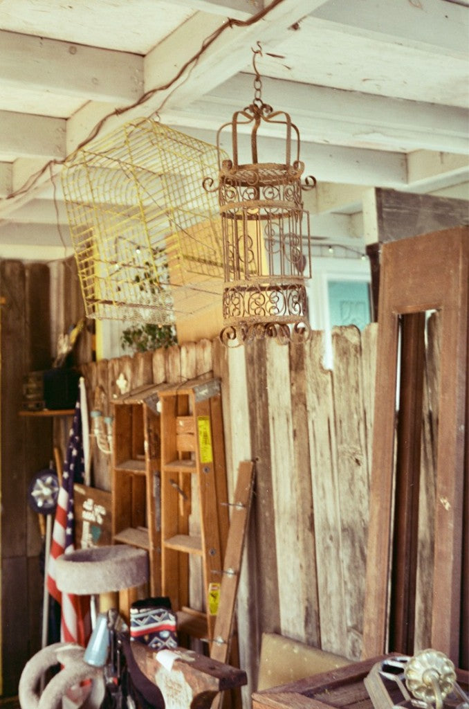 Covered antique porch with hanging birdcages and an iron lantern above a wooden fence, US flag, and ladder, shot on Amber 100 35mm film, developed in Kodak C-41