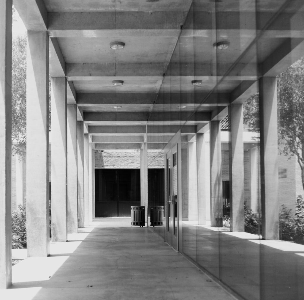 Covered concrete colonnade with glass-wall reflections and two trash cans at the far end, UTRGV School of Medicine campus, Edinburg, Texas, shot on Fujifilm Neopan Acros 100 II 120 film, developed in standard black-and-white chemistry