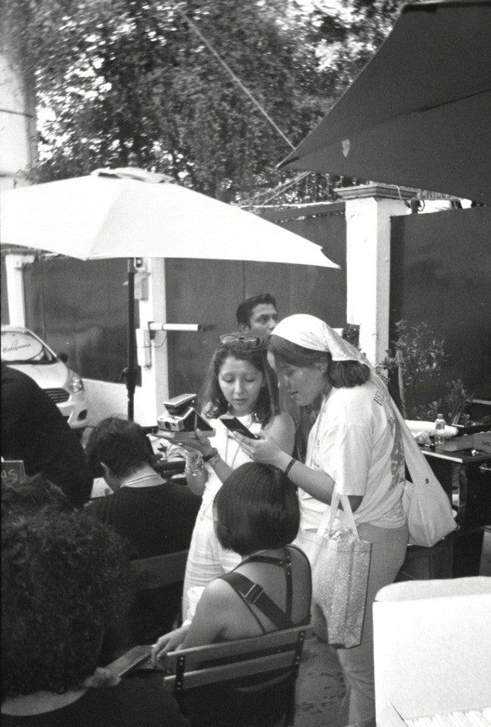 Two women at an outdoor café looking at a phone, one holding an instant camera, Kodak Ektar H35N + Ilford Delta 100 + 35mm half-frame