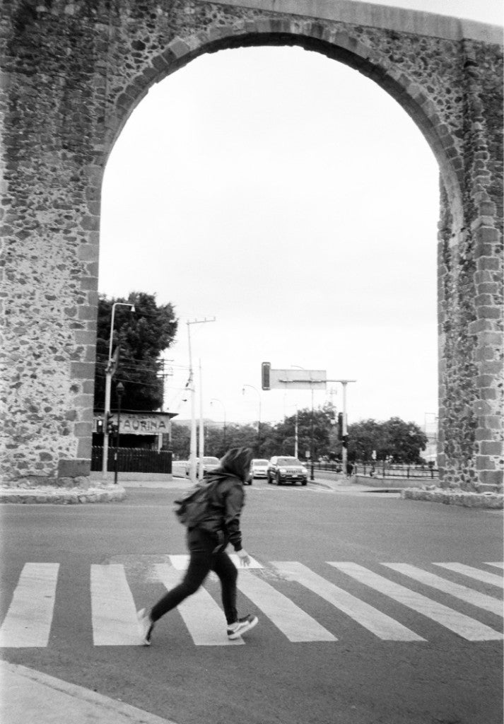 Pedestrian crossing under a stone aqueduct arch at Los Arcos de Querétaro, Calzada de los Arcos, Santiago de Querétaro, shot on Ilford HP5+ 35mm film, developed in standard black-and-white chemistry