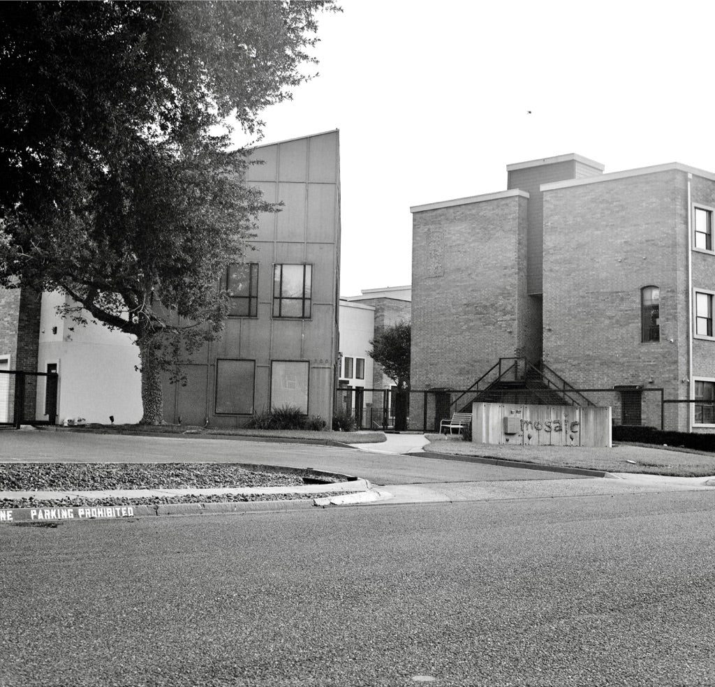 Mosaic apartments street view with mosaic monument sign, modern brick and metal buildings, and PARKING PROHIBITED curb, McAllen, shot on Ilford XP2 Super 120 film, developed in Kodak C-41
