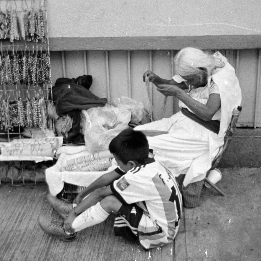 Elderly woman with long white hair threading beads beside a bracelet stand, a boy in a striped soccer jersey sitting on the sidewalk in front, Coyoacán, Mexico City — Kodak Tri-X 400 35mm disposable camera