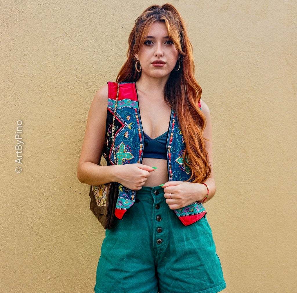 Woman with auburn hair in an embroidered vest and green shorts standing against a yellow wall shot on Lomography Metropolis 400 120 film, developed in C-41