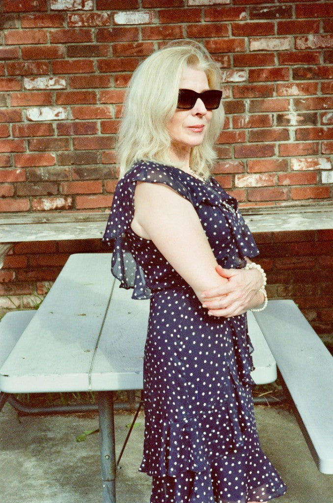 Mujer rubia con un vestido azul marino de lunares y gafas de sol de pie junto a una mesa de picnic contra una pared de ladrillos, fotografiada con película a color Kodak Vision 3 500T 5219 AHU, C-41) 35mm, revelada con revelador Kodak C-41.