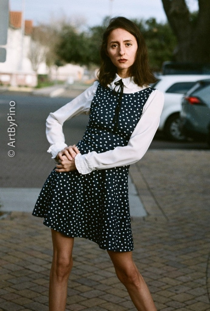 Fotografía de una mujer con un vestido azul marino de lunares sobre una blusa blanca, de pie en una acera con coches detrás, tomada con película de 50 fps y revelada con química estándar en blanco y negro.