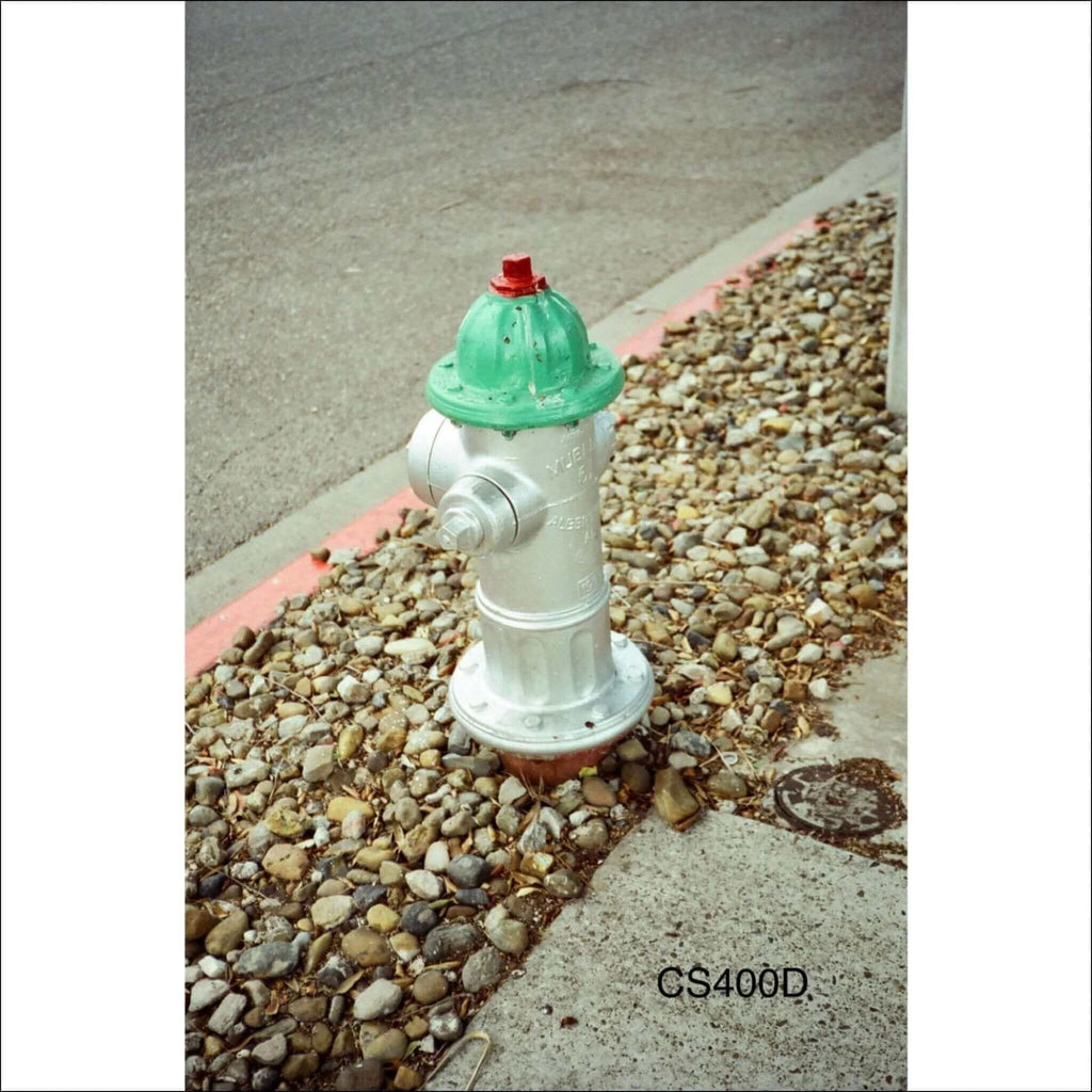 A silver fire hydrant with a green top and red cap, set in a gravel bed beside a sidewalk.