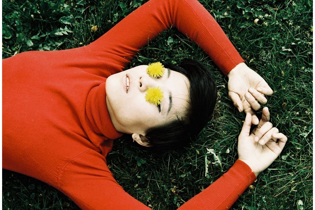 Person in red sweater lying on grass with yellow flowers over eyes, showcasing creative photography with color contrasts.