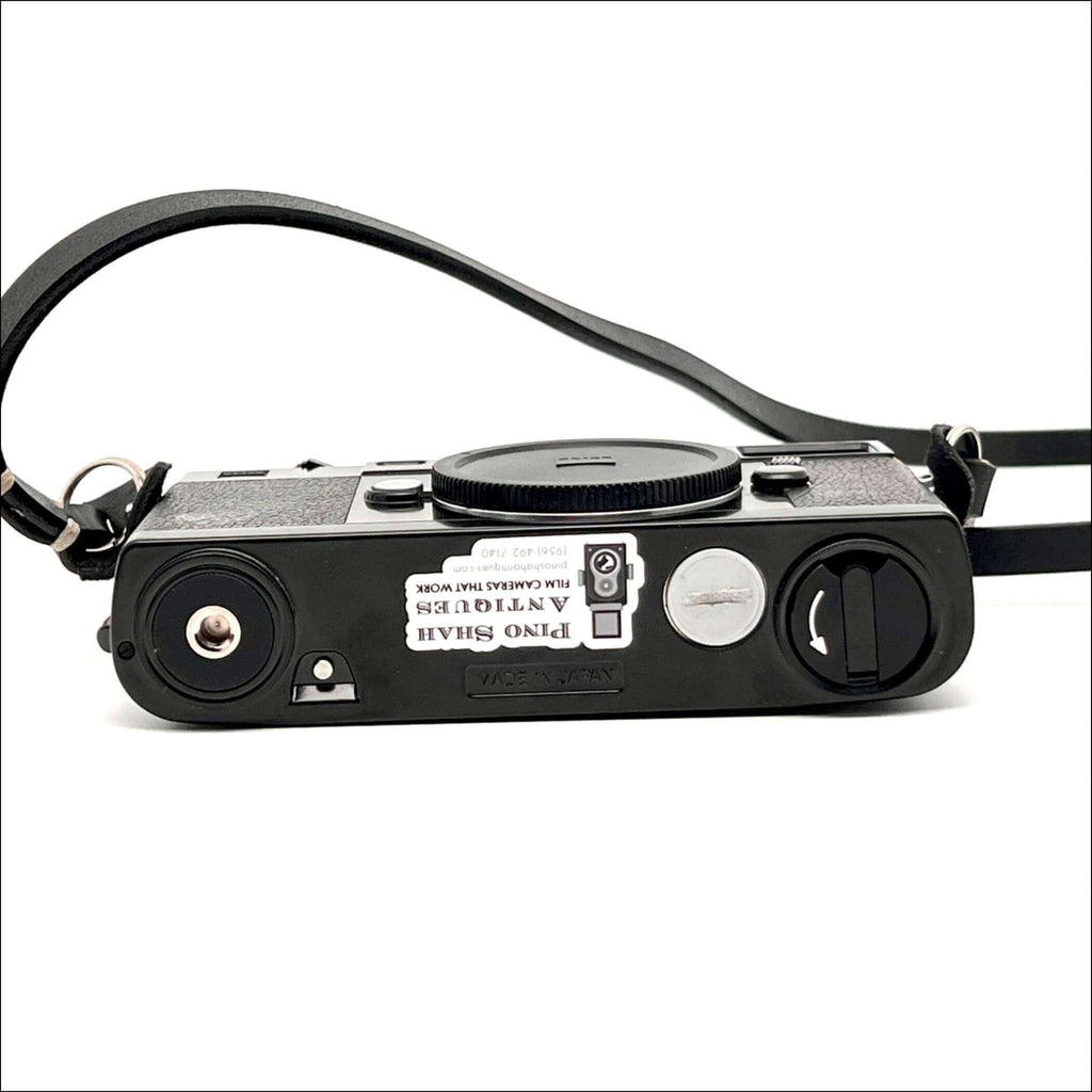 Black textured body with a silver shutter button and black leather strap, featuring a sticker that reads ’pino shah antiques film cameras that work.’