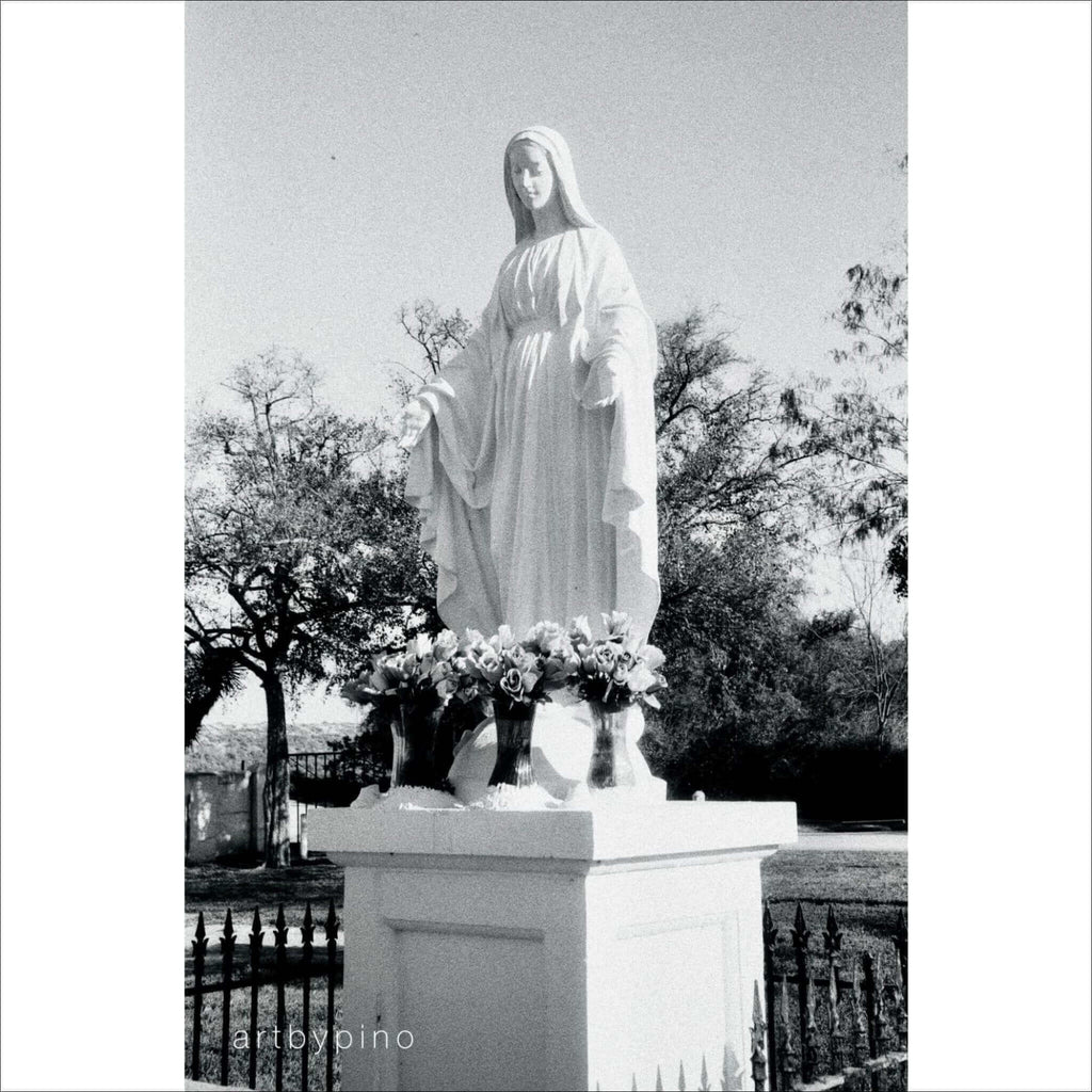 White marble statue of the virgin mary standing on a pedestal with flowers at its base.