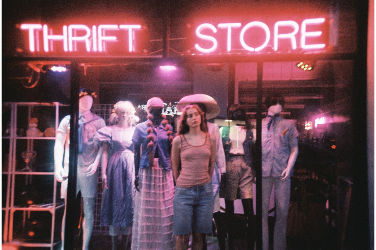A woman stands in front of a thrift store window display, wearing a tan tank top and blue denim shorts.