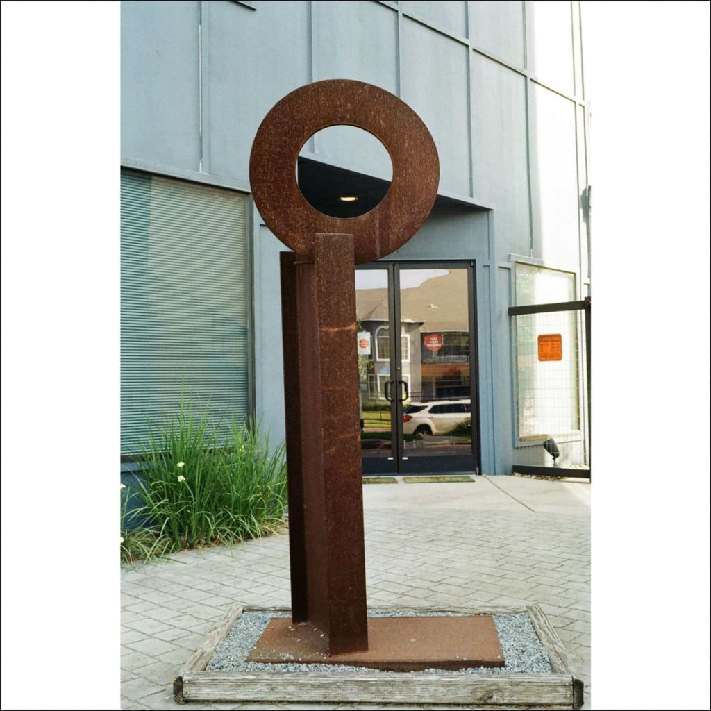 Rusty metal key-shaped sculpture with a circular head and vertical shaft, standing on a wooden base.