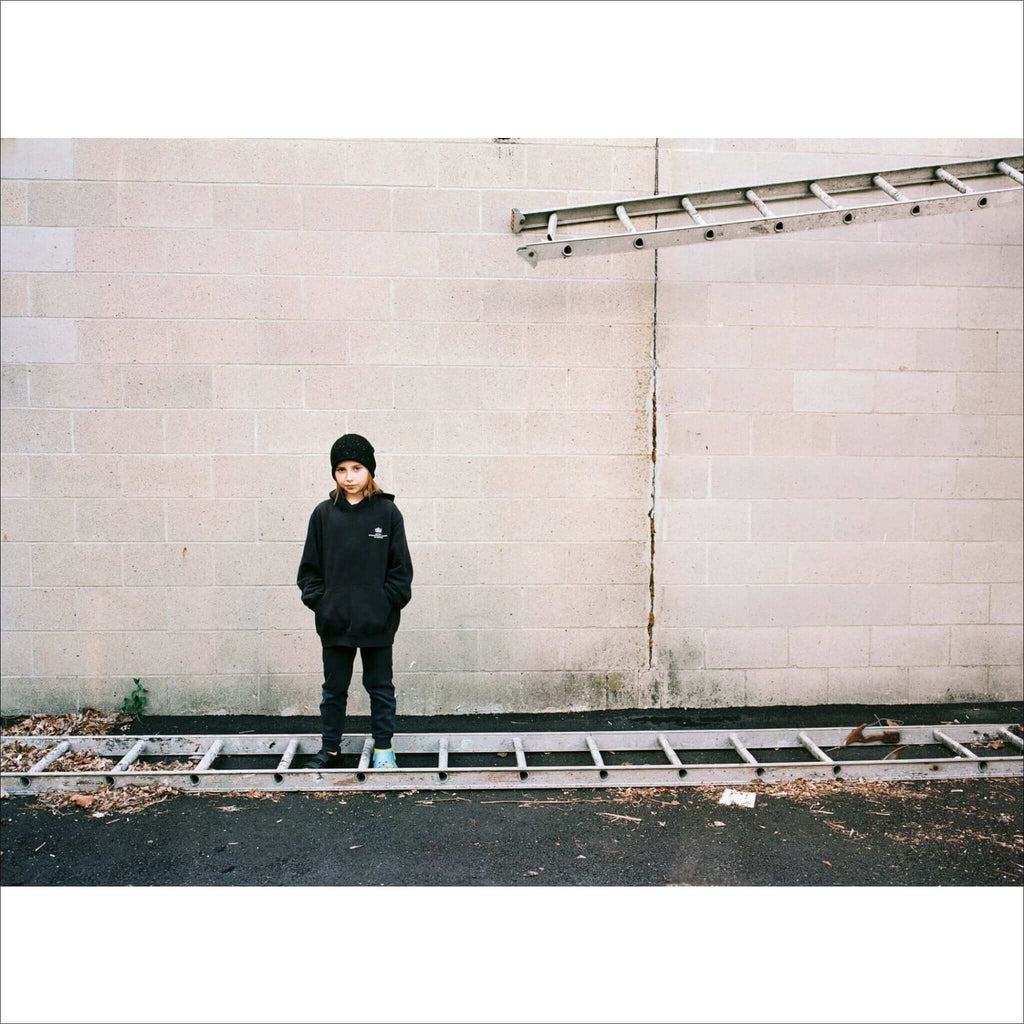 A child wearing a black hoodie and beanie stands against a concrete wall, with two silver ladders positioned horizontally—one leaning against the wall above them and another lying flat on the ground in front.