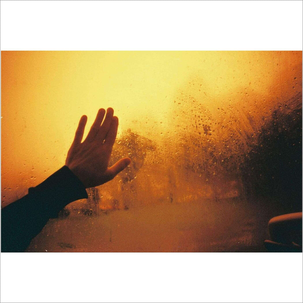 A hand pressed against a fogged-up window, fingers spread as if reaching out through the warm, golden haze.