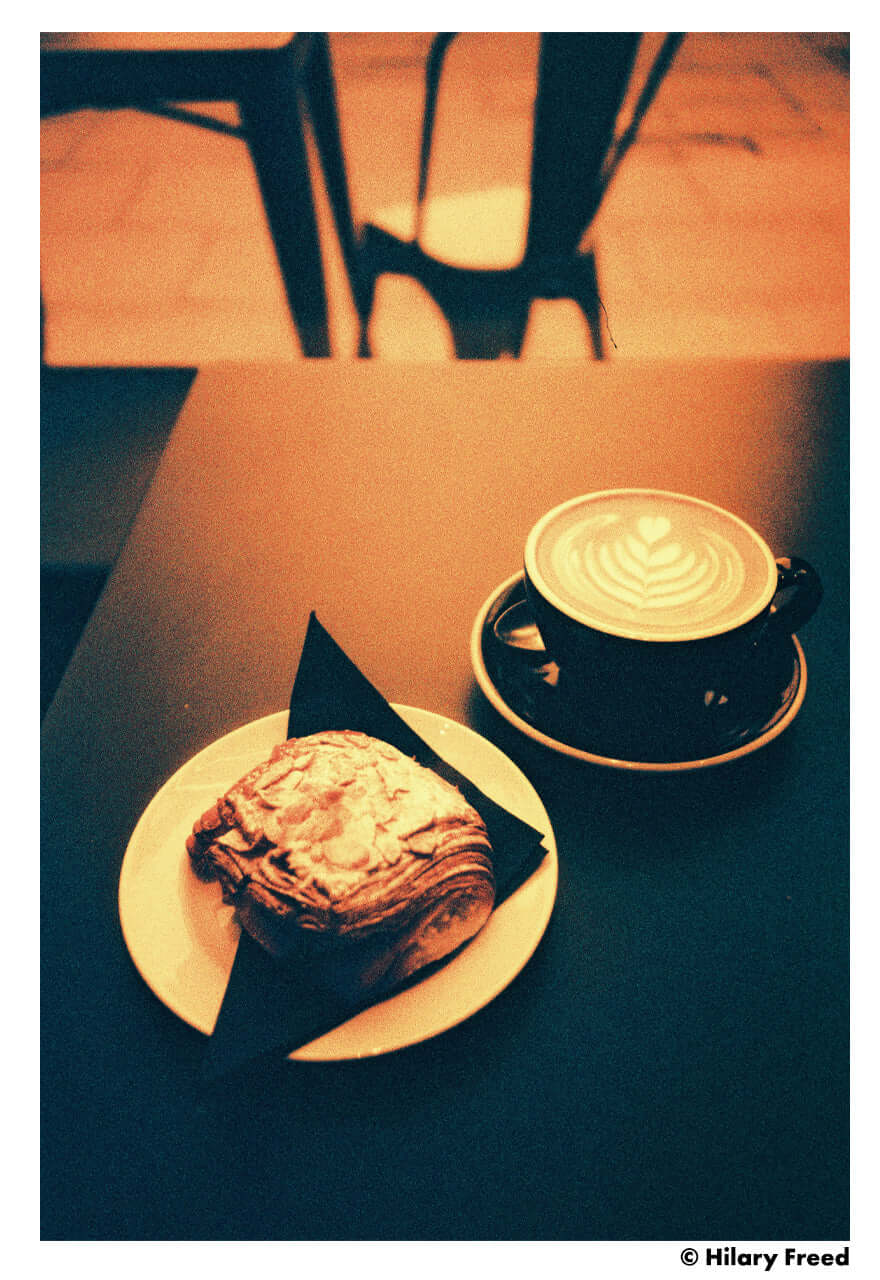 A delectable pastry on a plate with a coffee cup featuring latte art on a dark table.