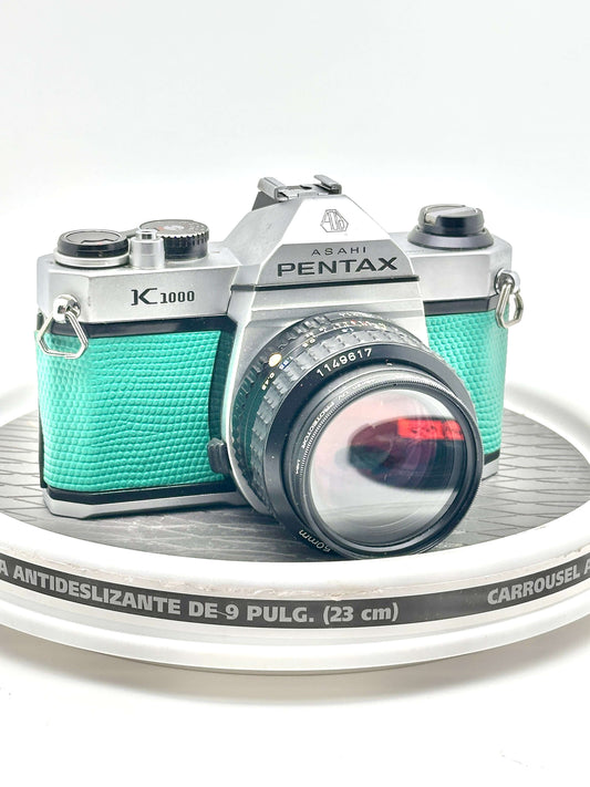 Japanese Asahi Pentax K1000 teal 35mm film camera with 50mm f1.4 lens on display, showcasing its classic design.