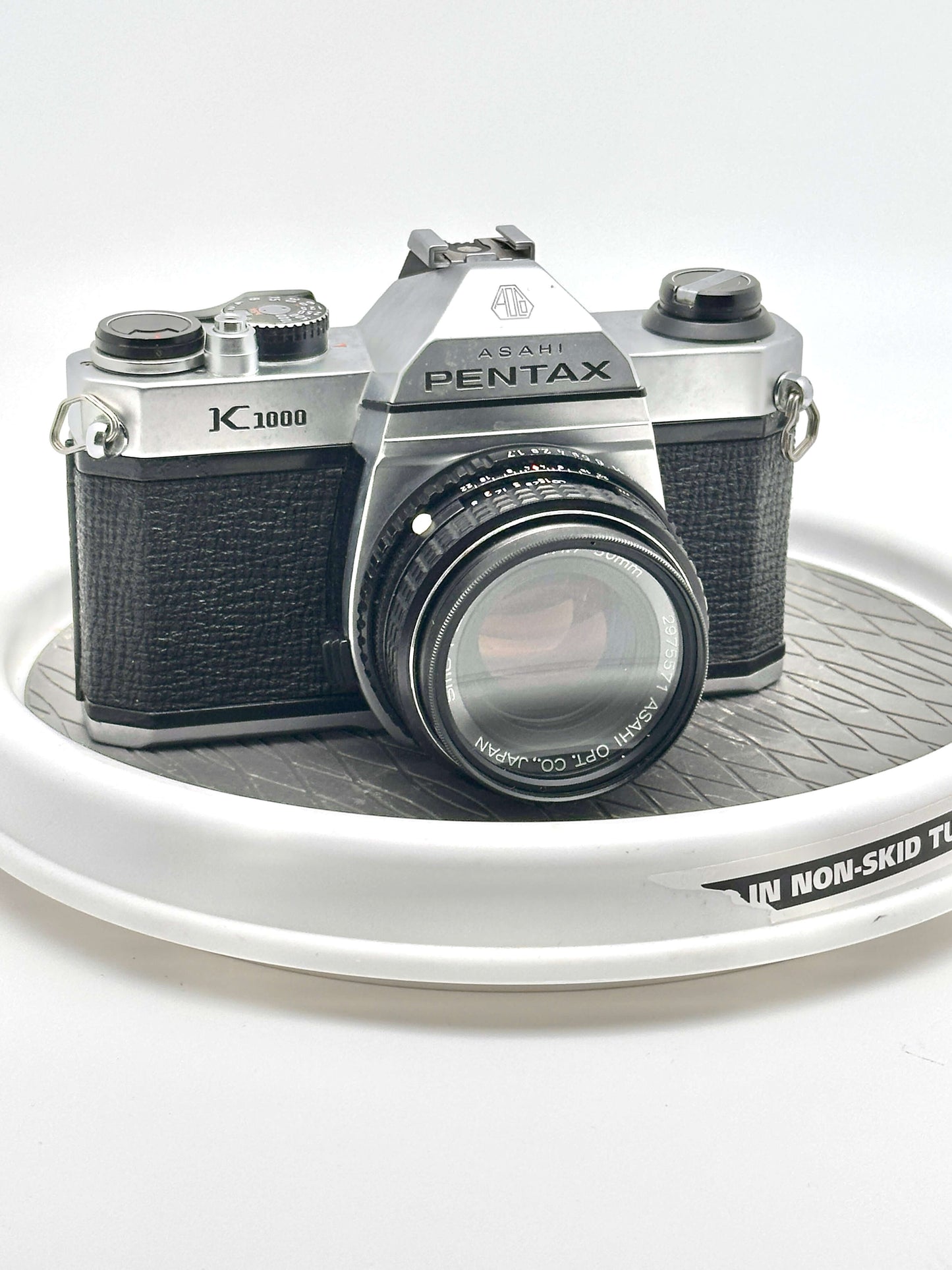 Japanese Asahi Pentax K1000 Black 35mm film camera with 50mm f1.7 lens displayed on a white circular tray.