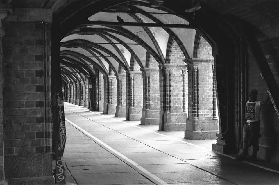 Black and white photo of a brick arcade with arched columns and a person leaning against the wall.