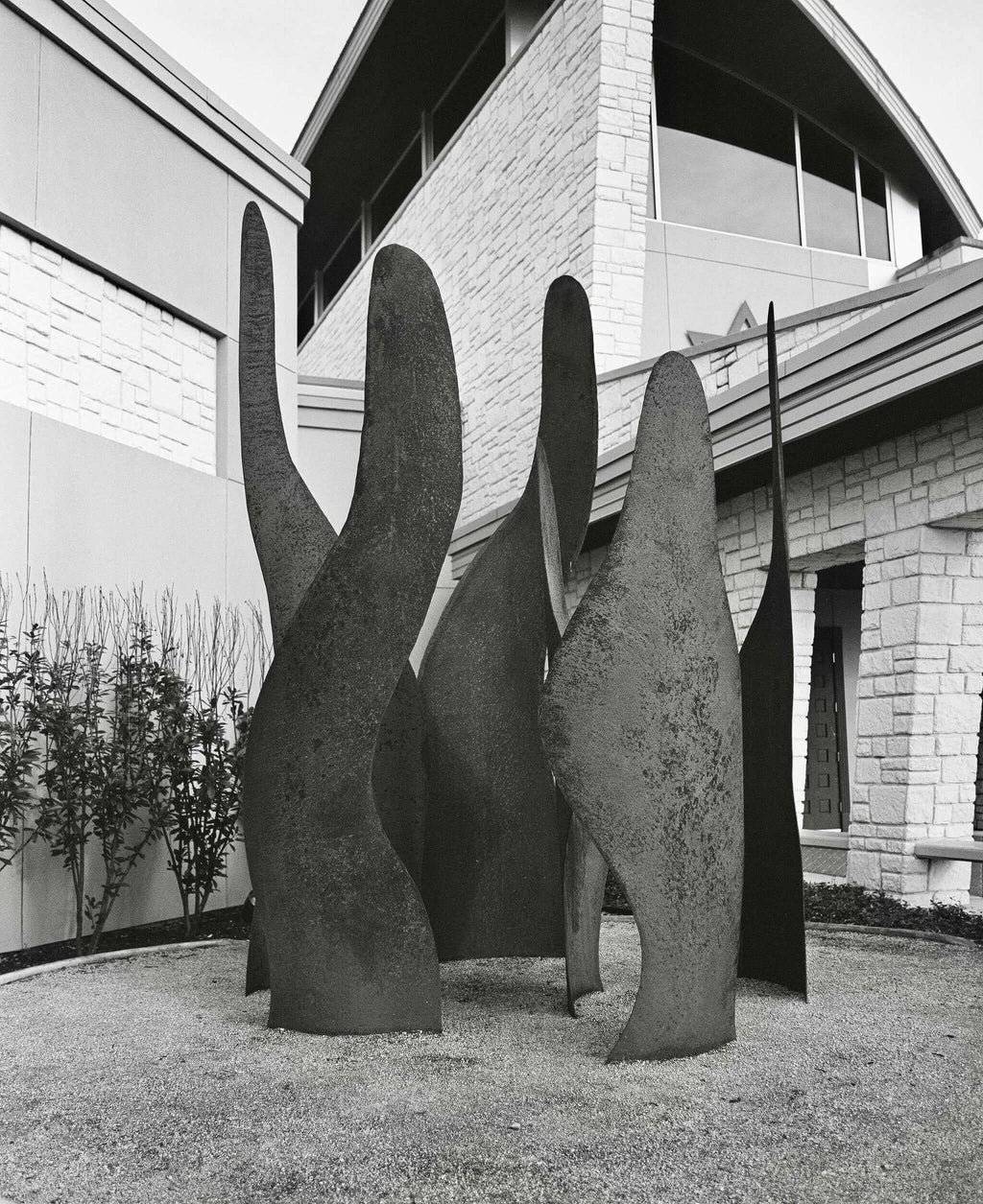 Black and white photograph of abstract metal sculptures in a landscaped area near a modern building.
