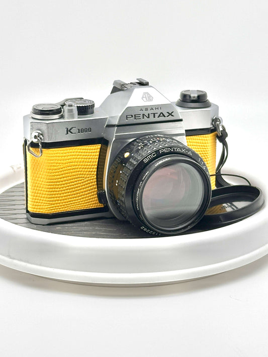 Japanese Asahi Pentax K1000 yellow 35mm film camera with 50mm f1.4 lens on a white turntable.
