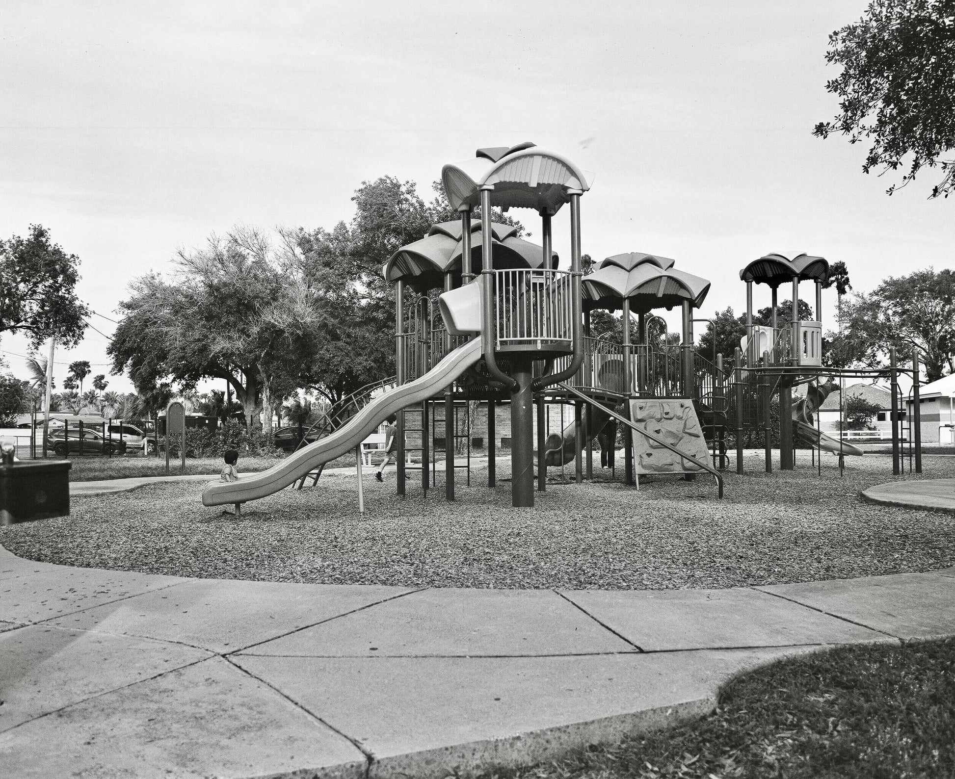 Black and white photo of a playground featuring slides and climbing structures surrounded by trees.