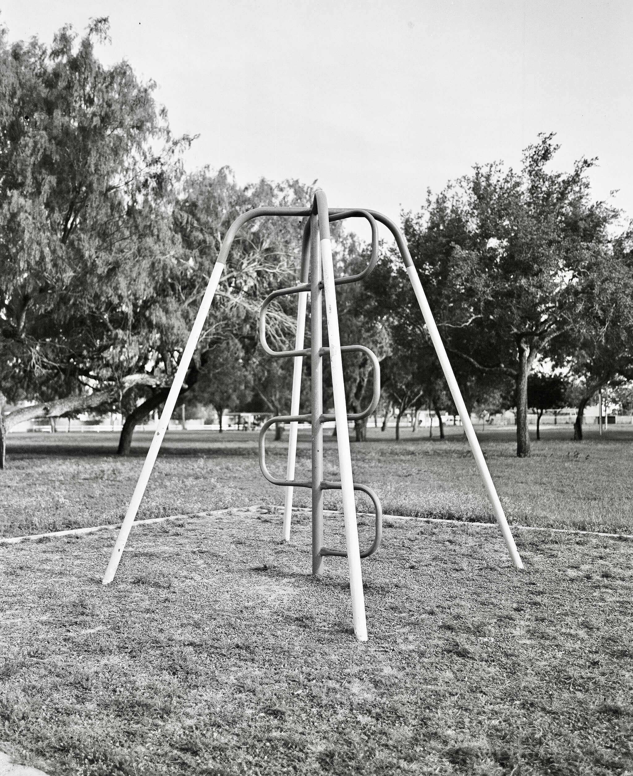 Black and white photograph of a playground swing set surrounded by trees in a grassy park.