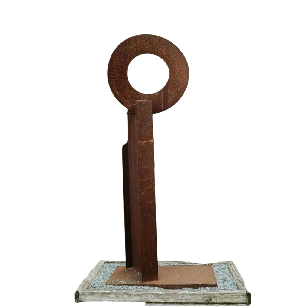 Rusty metal key-shaped sculpture with a circular head and vertical shaft, standing on a wooden base.