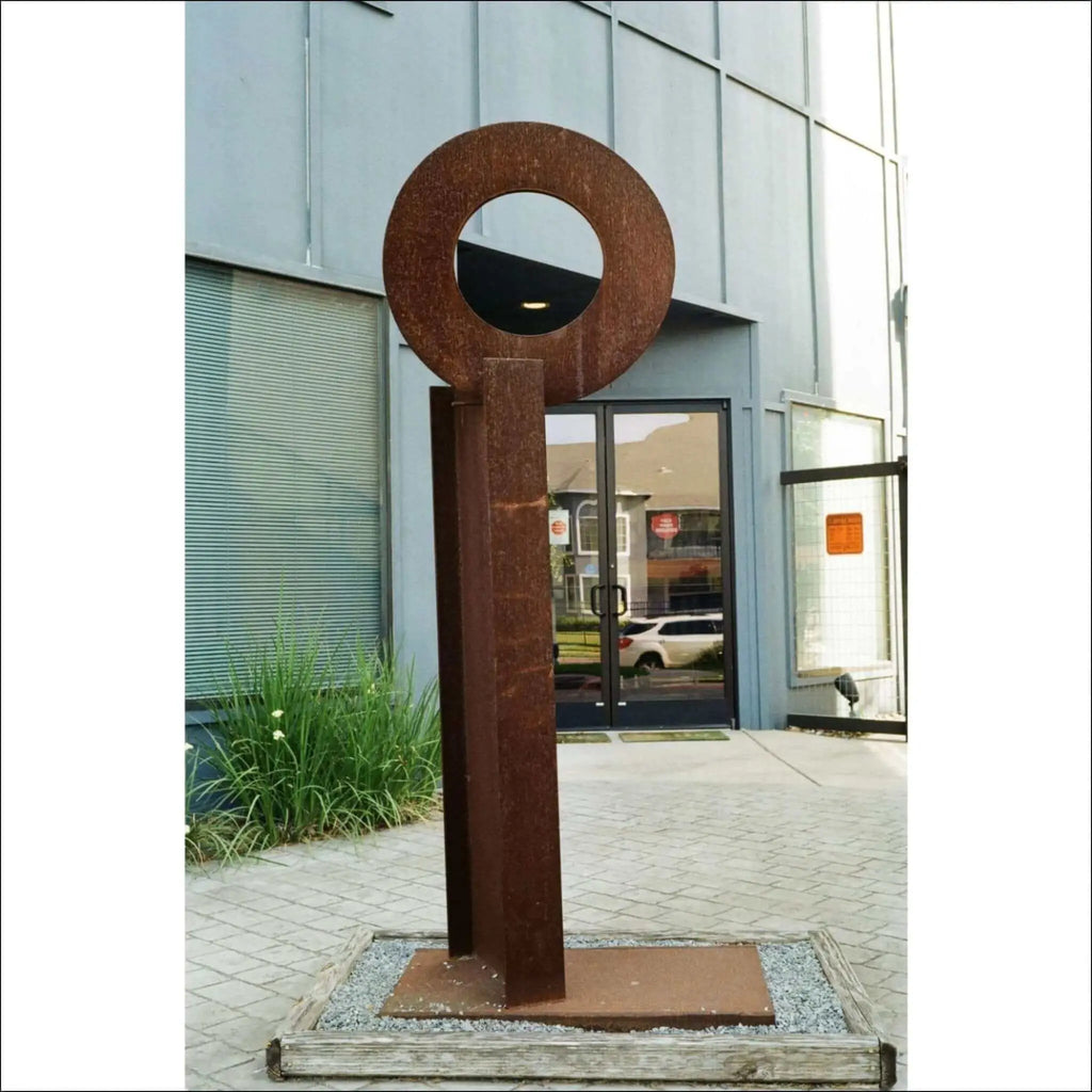 Rusty metal key-shaped sculpture with a circular head and vertical shaft, standing on a wooden base.