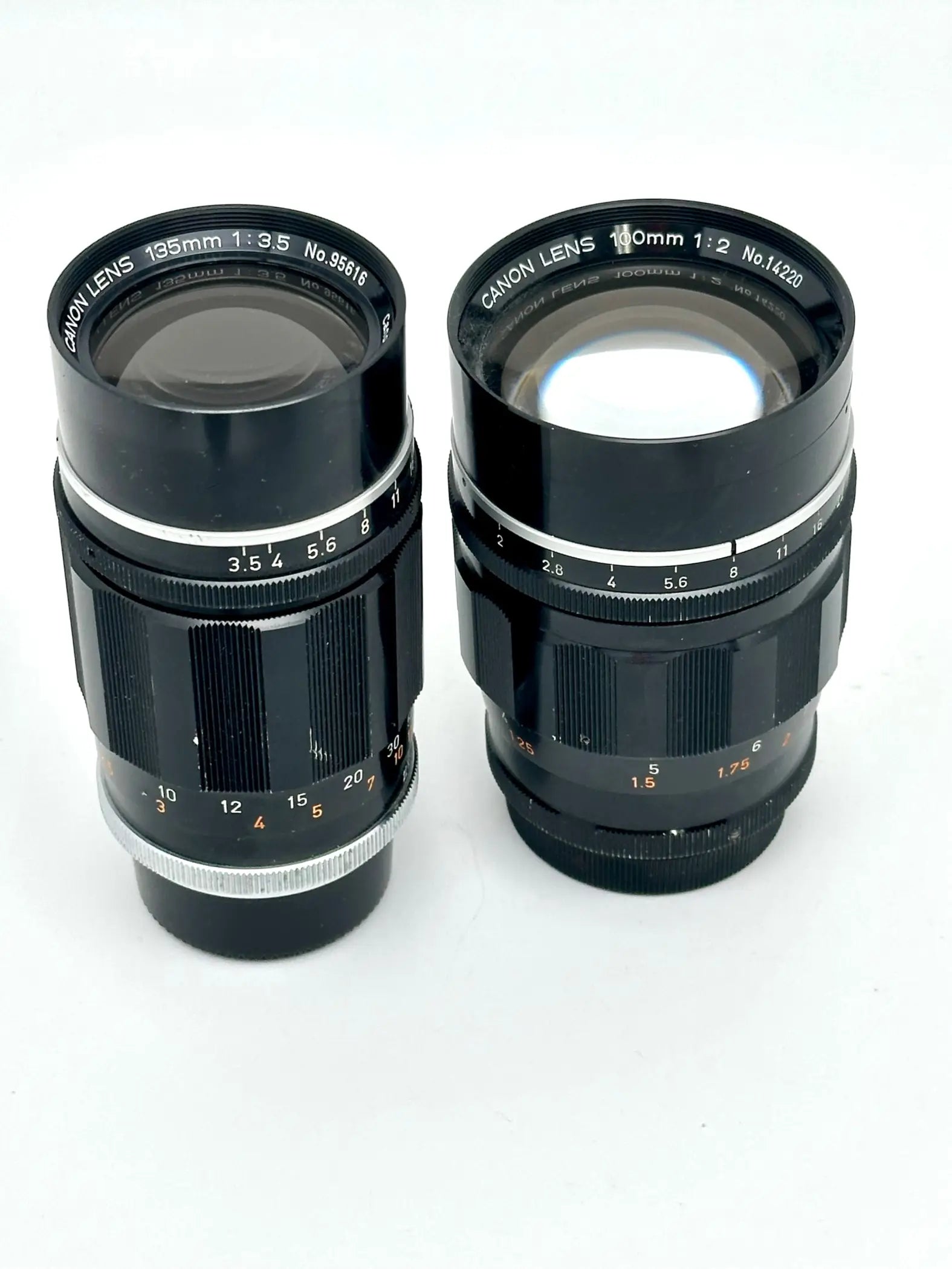 Two black canon camera lenses with silver metallic rings and engraved markings, one labeled 195mm f/3.5 and the other 100mm f/2.