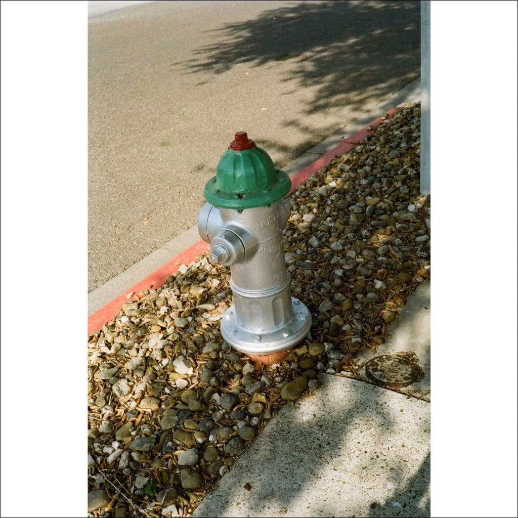 A silver fire hydrant with a green top and red cap, set in a gravel bed beside a sidewalk.
