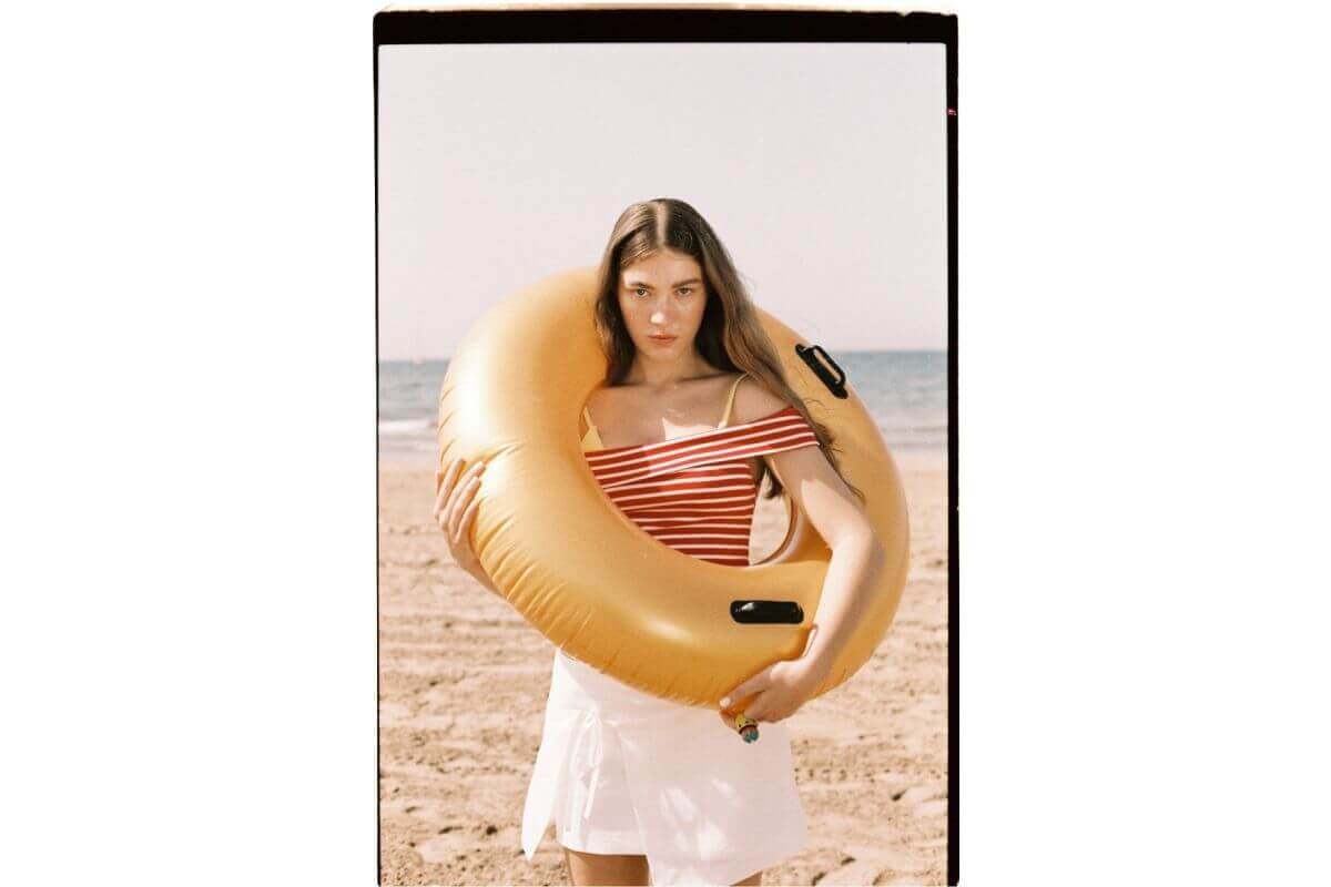 Woman in striped swimsuit holding a gold inflatable ring on a sunny beach.