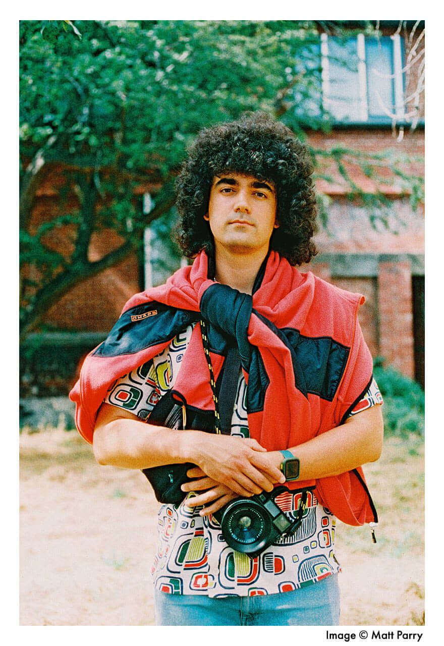 Young man with curly hair and colorful shirt holding a camera, standing outdoors with trees in the background.