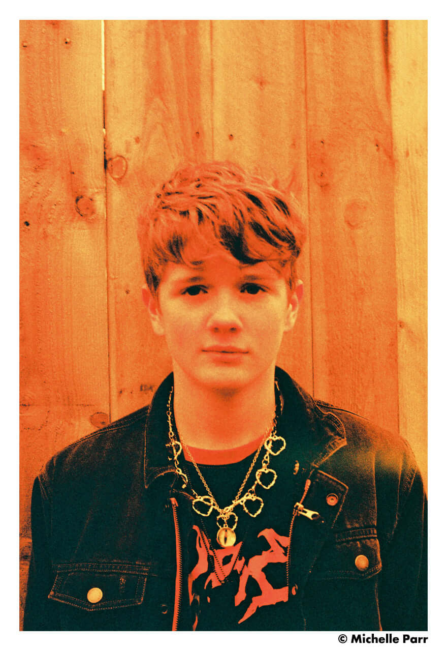 Portrait of a young person with short hair, wearing a black jacket and jewelry, against a wooden background.