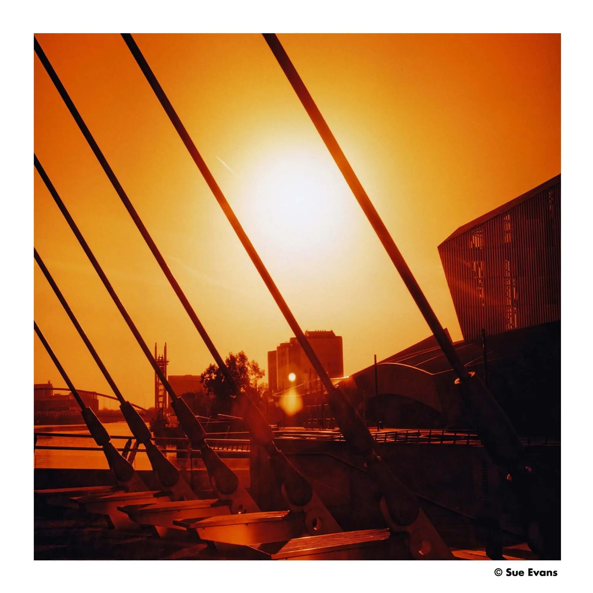 A modern bridge with sleek, dark metal cables stretching diagonally across a warm orange sunset sky.