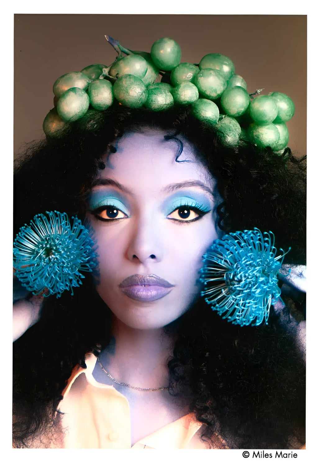A woman wears a green grape crown and blue dandelion-like earrings.