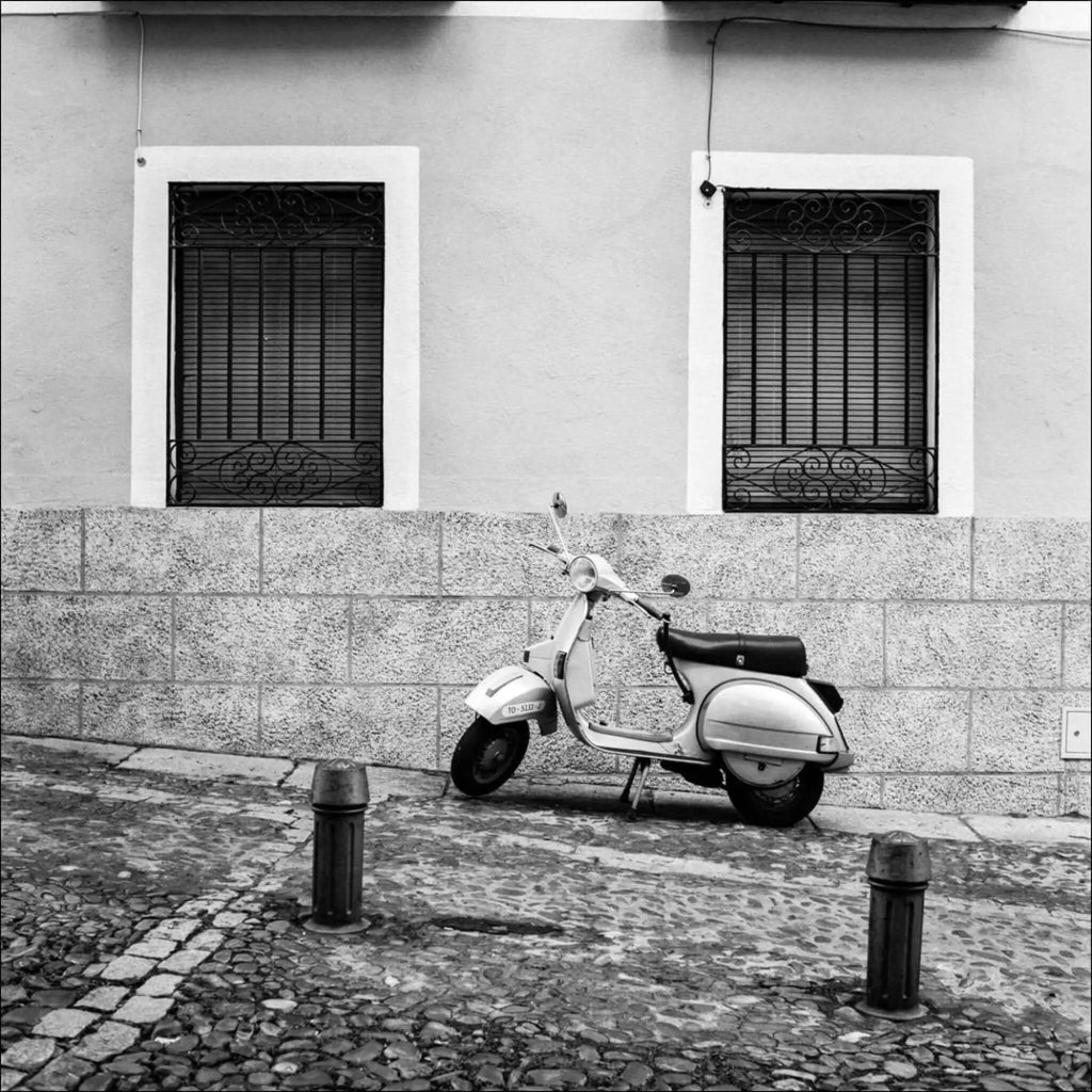A classic white vespa scooter with a black seat, parked against a stone wall.