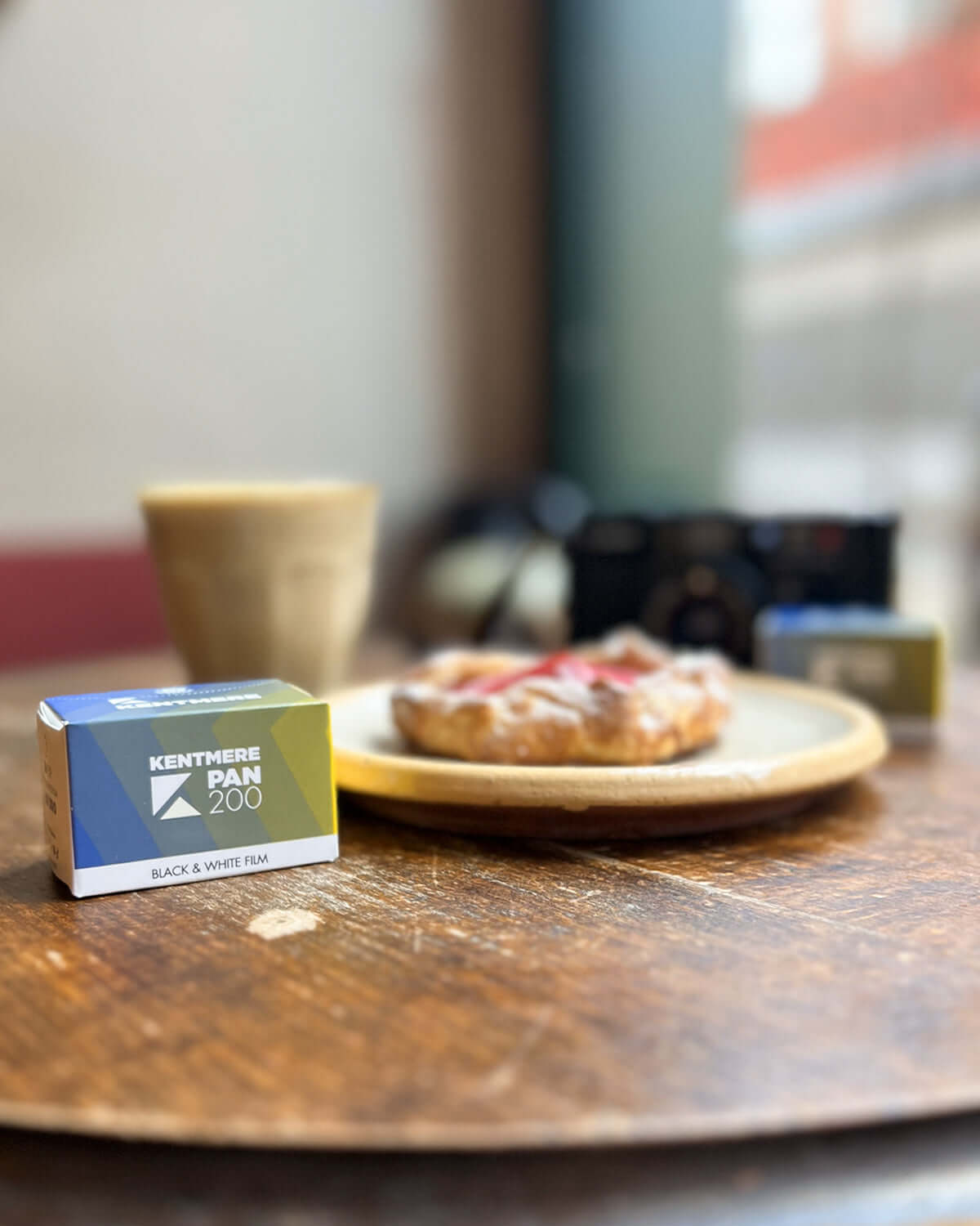 Kentmere Pan 200 black and white film displayed on a wooden table with a pastry and coffee cup.