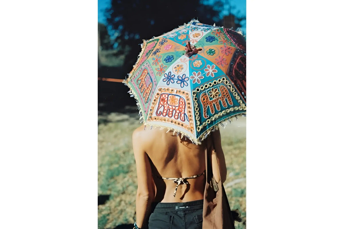 A vibrant, hand-painted indian-style umbrella with intricate floral and elephant motifs in turquoise, orange, red, and blue hues, adorned with decorative fringe along its edges.
