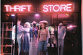 A woman stands in front of a thrift store window display, wearing a tan tank top and blue denim shorts.