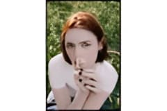 A woman with reddish-brown hair, wearing a white long-sleeved top and holding her hands near her face.