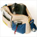 Beige canvas shoulder bag with blue grid-patterned pocket and yellow zipper pulls, holding a black polaroid camera.