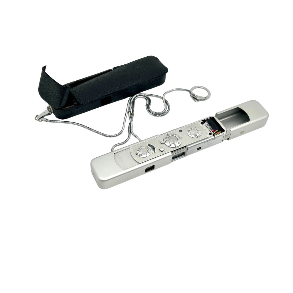 Silver metallic compact camera with circular dials and a retractable lens, accompanied by its black leather case.