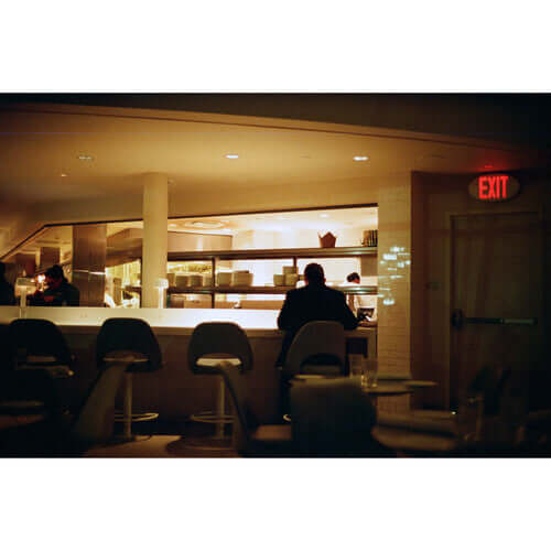 Dimly lit restaurant interior featuring bar seating and chefs at work in the background.
