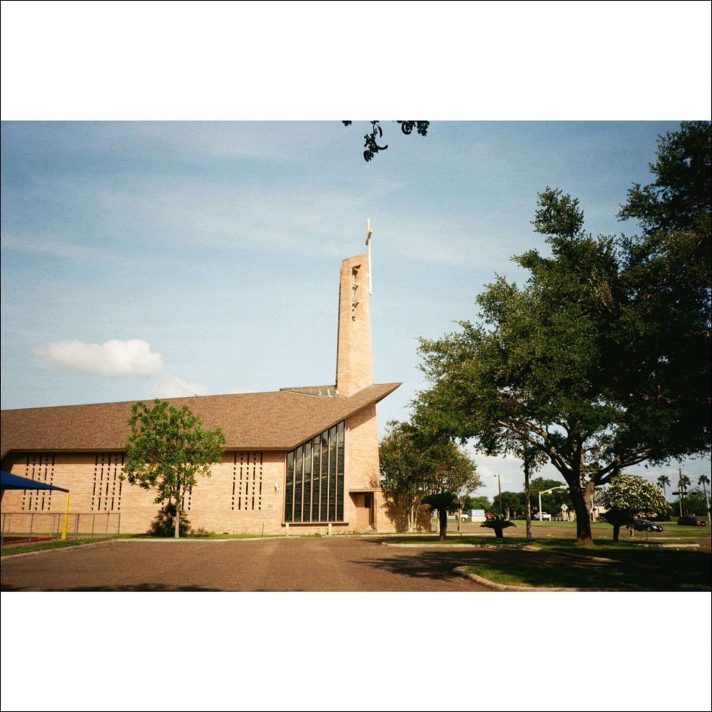 Beige stone church with a tall, slender tower topped by a cross and large glass windows.