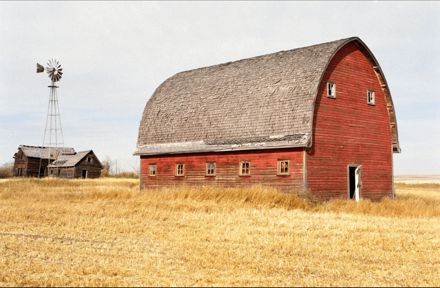 Rustic red barn in golden field with a windmill and old shed in the background on a cloudy day.