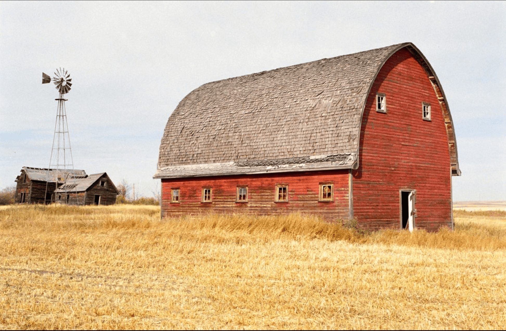 Rustic red barn in golden field with a windmill and old shed in the background on a cloudy day.