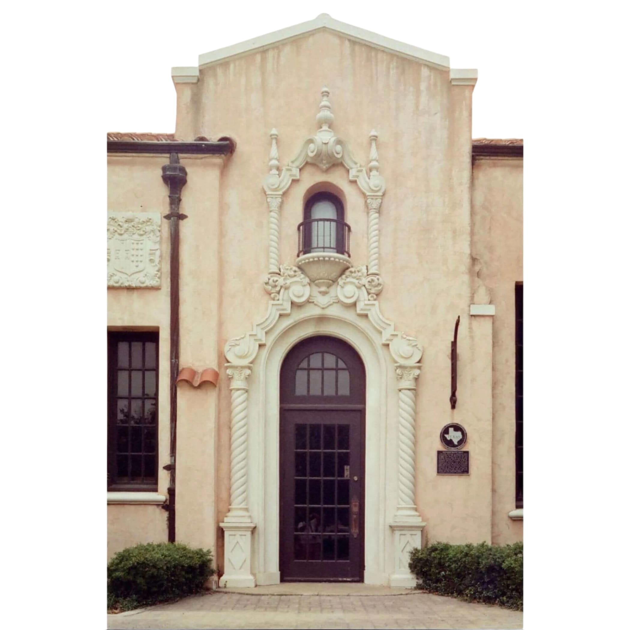The image shows a beige stucco building with an ornate white stone archway framing a dark brown arched door.